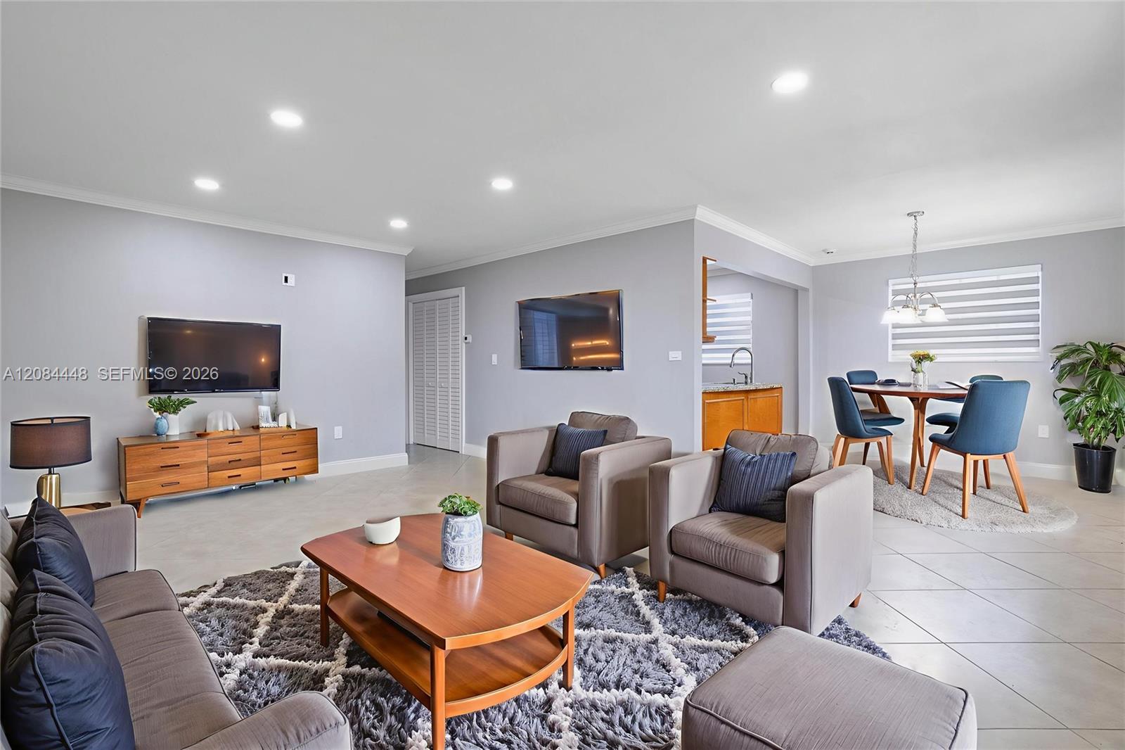 Step into an airy and open living space filled with natural light and timeless charm. The living room features neutral tile flooring, soft lavender-gray walls, and elegant crown molding for a polished finish. Recessed LED lighting enhances the fresh and m