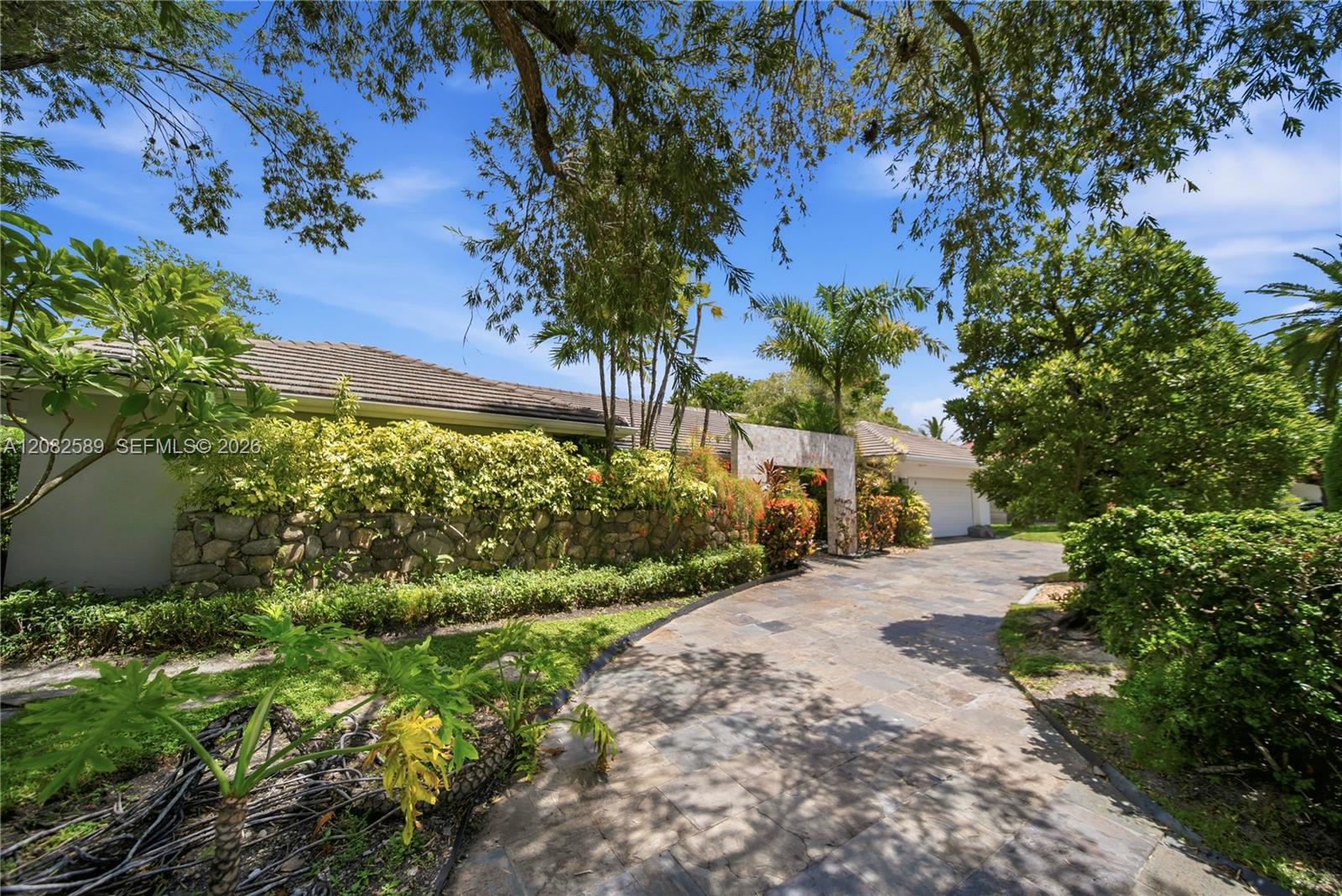 Front exterior featuring a spacious paved driveway, lush tropical landscaping, mature trees, and a private, inviting entrance.